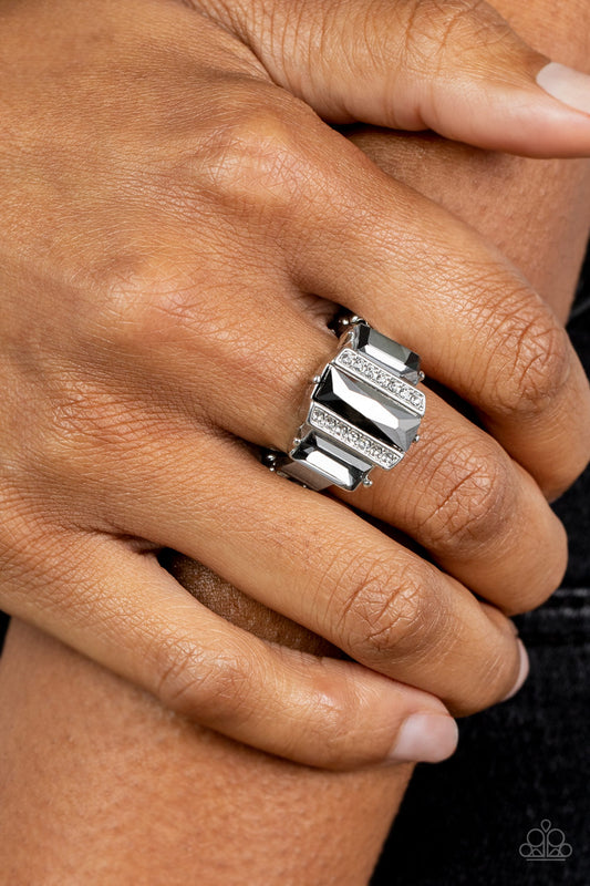 Paparazzi Accessories - A Glitzy Verdict - Silver Ring bordered by two rows of dainty white rhinestones, an emerald cut hematite rhinestone adorns the center of a band dotted with a pair of smoky emerald cut rhinestones, resulting into a jaw-dropping dazzle across the finger. Features a stretchy band for a flexible fit.  Sold as one individual ring.