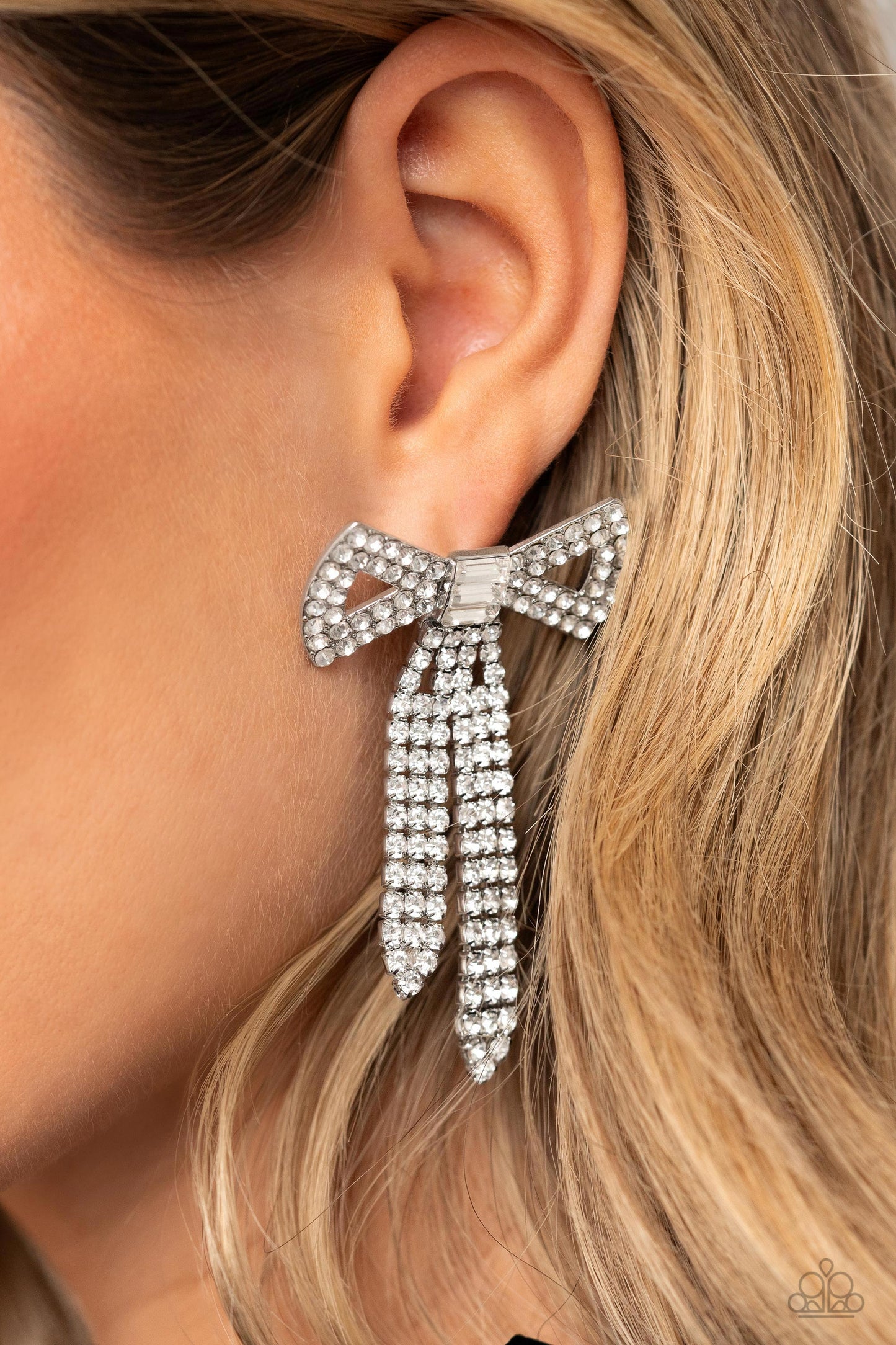 Paparazzi Accessories - Just BOW With It - White Earrings featuring silver square fittings, high-sheen bands of silver, adorned in sparkling white rhinestones loop into a stunning bow charm, creating a classy statement at the ear. Featured in the center of the classy design, a trio of emerald-cut white gems tie the bow together emitting further sheen and glitz. Earring attaches to a standard post fitting. Sold as one pair of post earrings.