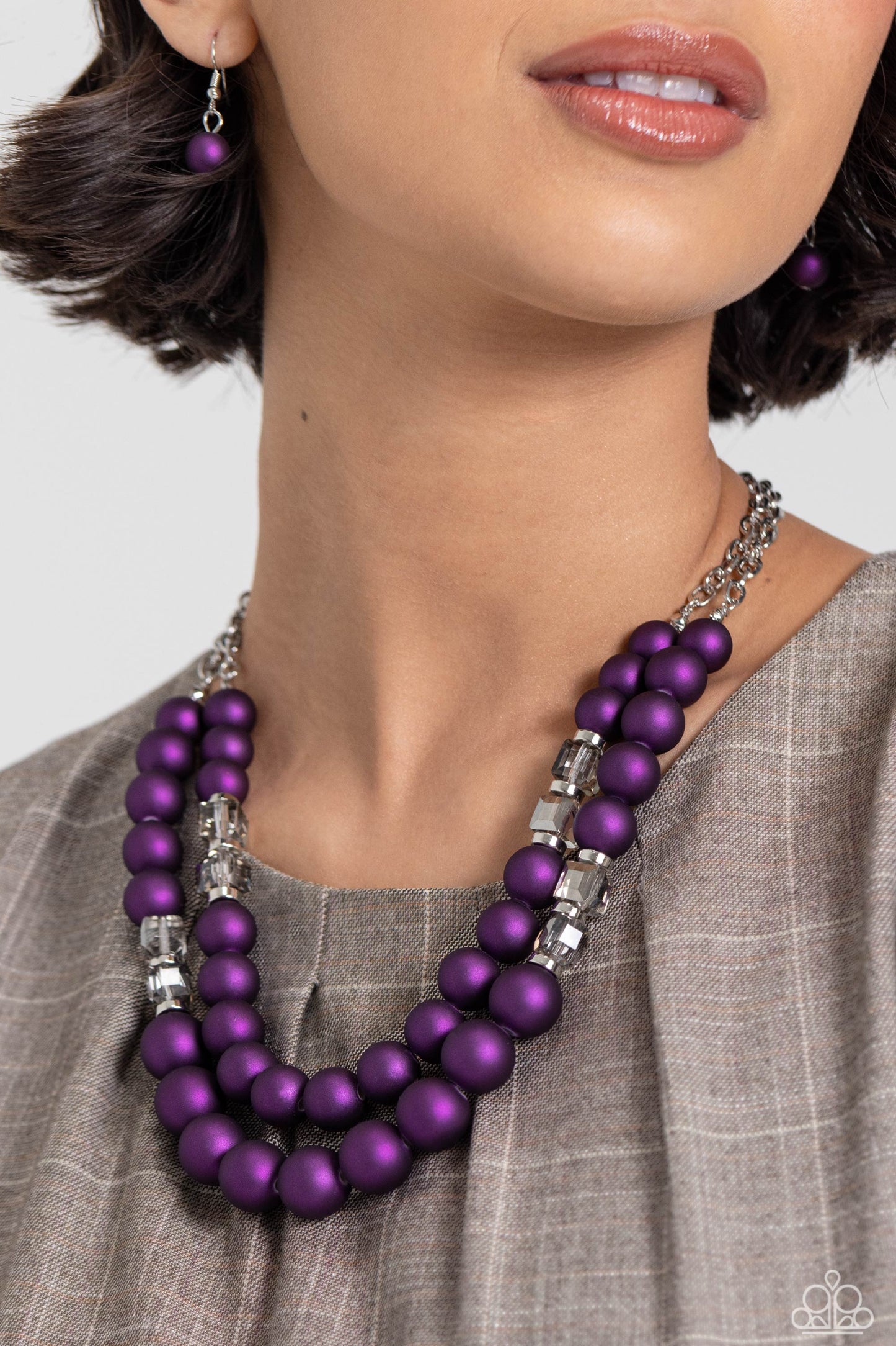 two rows of oversized purple beads featuring a subtle shimmer, silver accents, and clear gray cubed beads fall below the collar on silver paperclip chains, creating refined, colorful layers. Features an adjustable clasp closure. Sold as one individual necklace. Includes one pair of matching earrings. Two strands of oversized purple beads featuring a subtle shimmer, silver accents, and clear gray cubed beads stretch around the wrist, creating refined, colorful layers.
