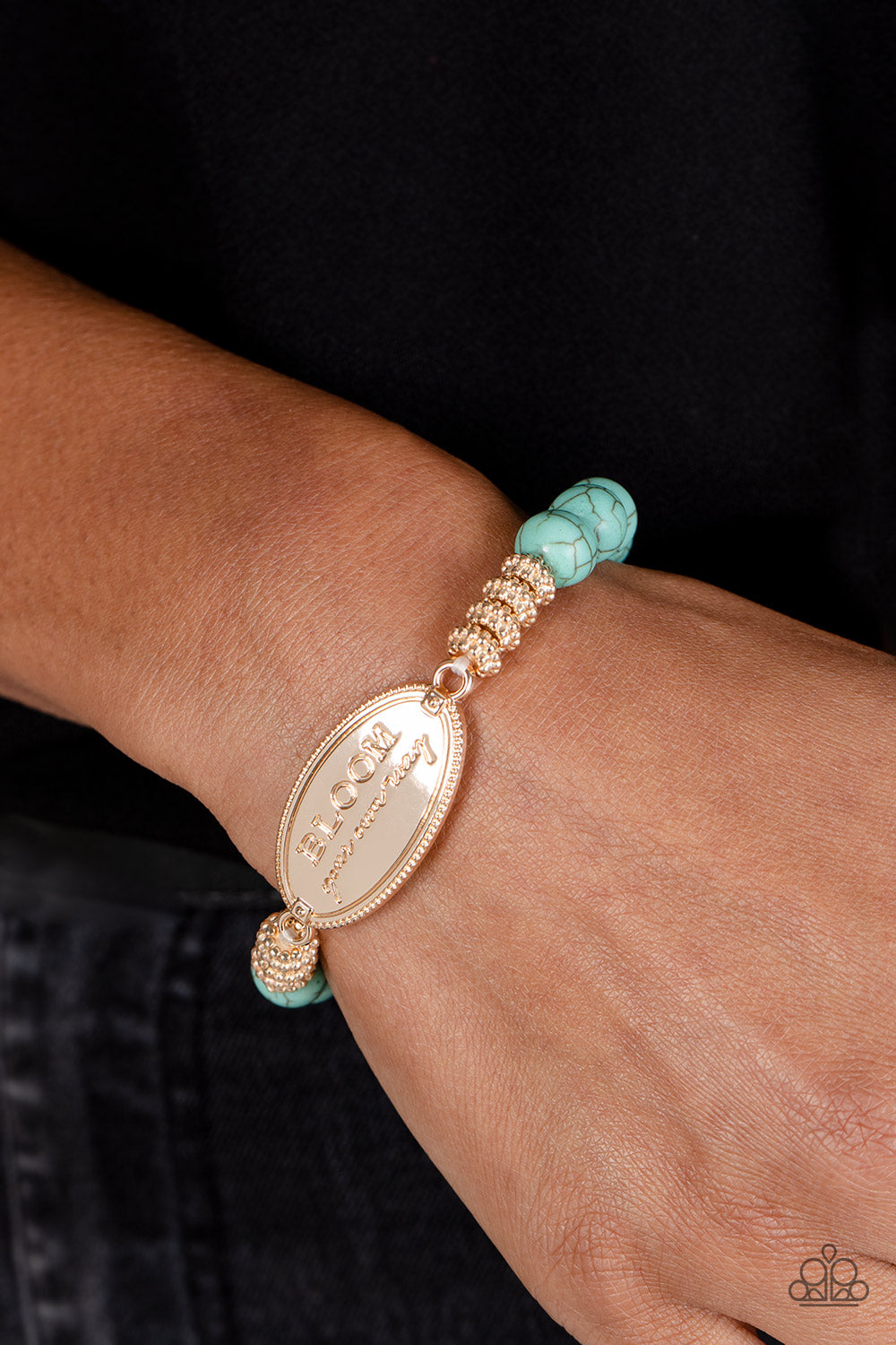 An asymmetrical, textured, gold oval pendant features the phrase "BLOOM your own way" in a variety of fonts to emphasize the inspirational message. Four textured gold wheels fan out from the pendant to a refreshing collection of light blue stones that stretch along the wrist on an elastic stretchy band for a charming pop of color along the wrist. As the stone elements in this piece are natural, some color variation is normal.