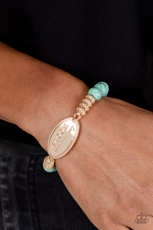 An asymmetrical, textured, gold oval pendant features the phrase "BLOOM your own way" in a variety of fonts to emphasize the inspirational message. Four textured gold wheels fan out from the pendant to a refreshing collection of light blue stones that stretch along the wrist on an elastic stretchy band for a charming pop of color along the wrist. As the stone elements in this piece are natural, some color variation is normal.