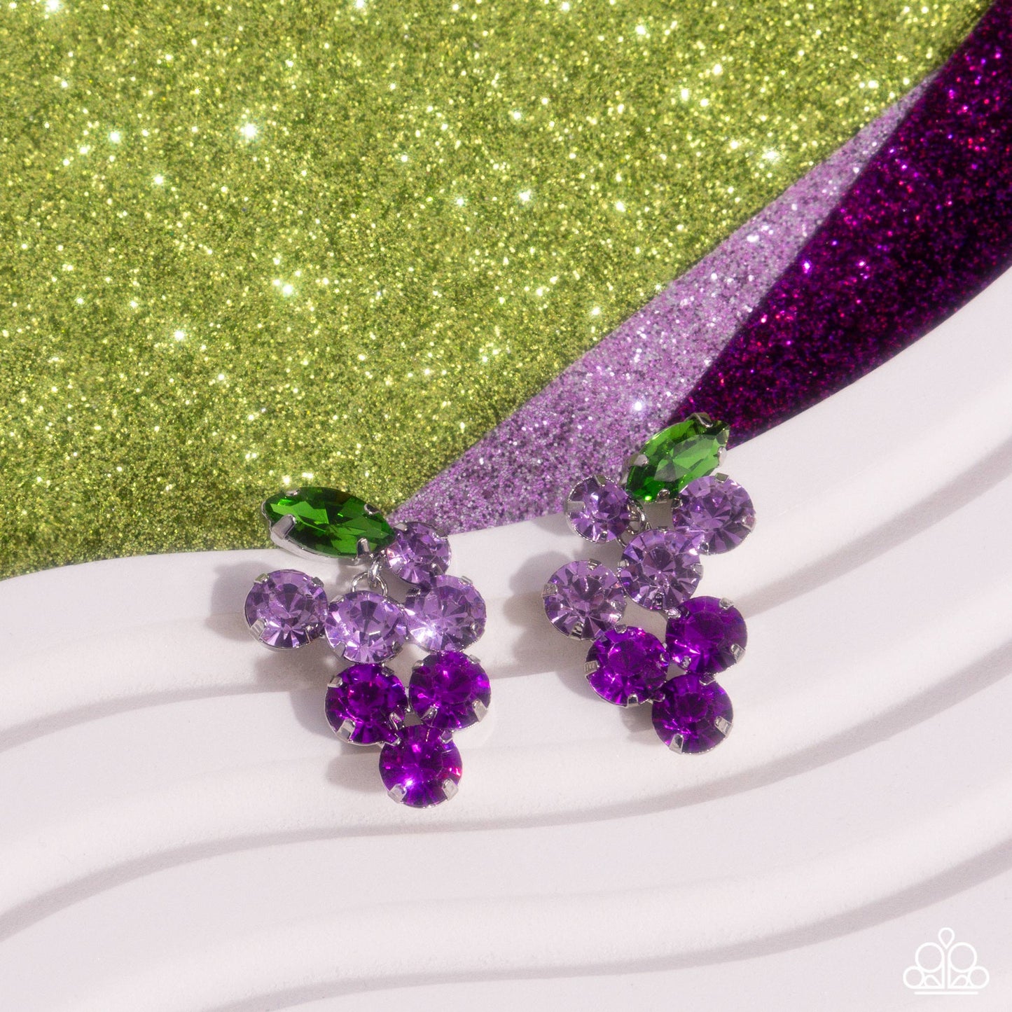 Pronged in silver fittings, a collection of round light purple rhinestones fade to darker purple rhinestones with a solitaire green marquise-cut leaf for an ombré, glittery grape-inspired display. Earring attaches to a standard post fitting.  Sold as one pair of post earrings.