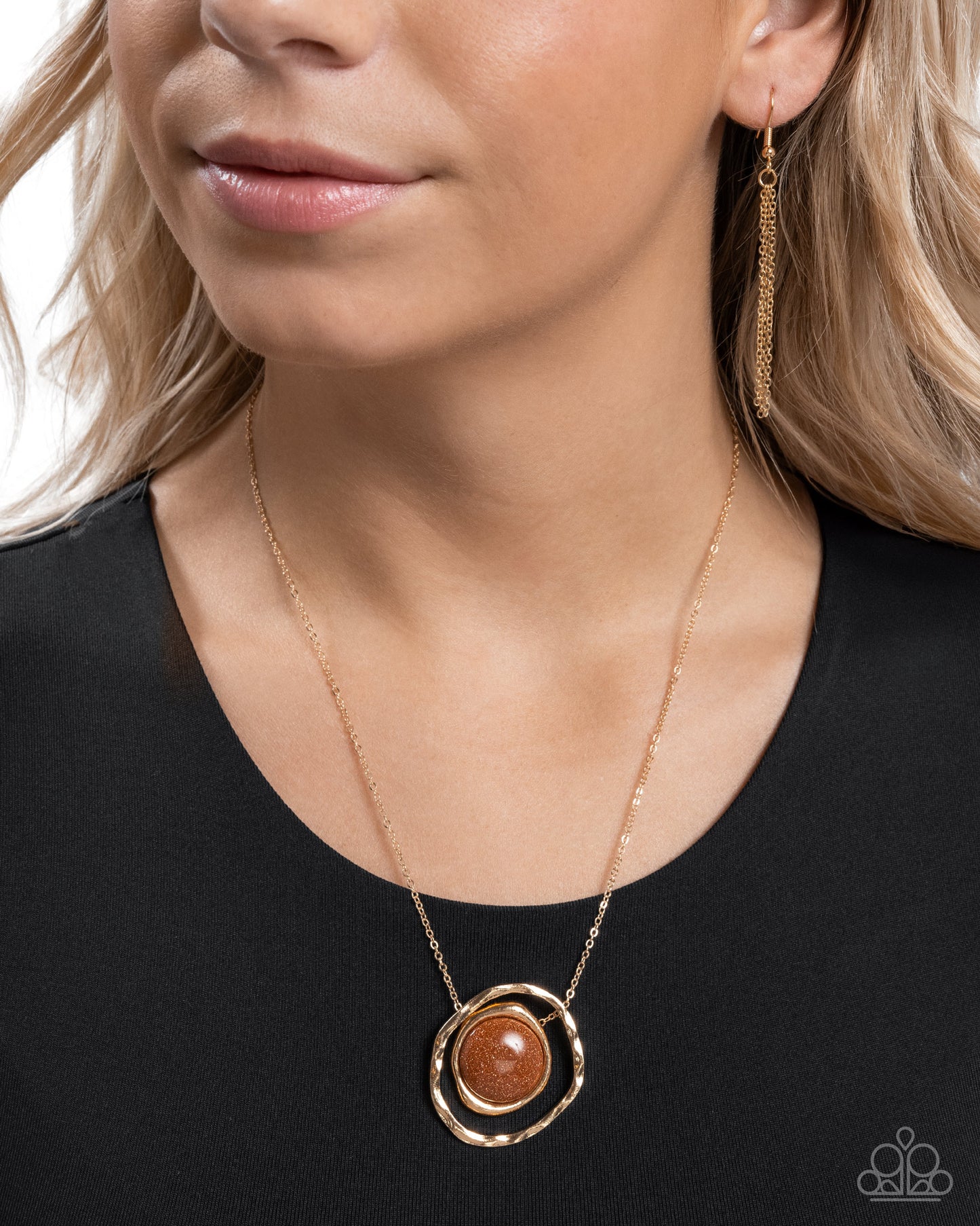 Suspended along the center of an oversized, warped, hammered gold hoop, a goldstone pendant, pressed in a smaller warped gold frame glides along a dainty gold chain for a glamorously gentle look. Features an adjustable clasp closure. As the stone elements in this piece are natural, some color variation is normal.  Sold as one individual necklace. Includes one pair of matching earrings.