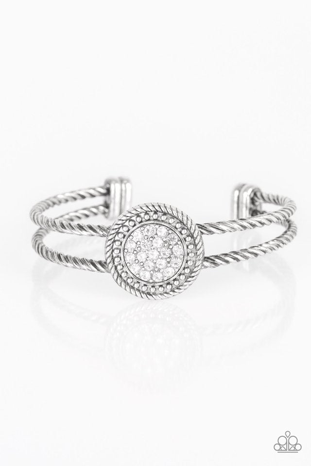 Paparazzi Definitely Dazzling - Silver Cuff Bracelets brushed in an antiqued shimmer, twisted silver bars arc across the finger, coalescing into an airy cuff. Radiating in silver studs and glittery white rhinestones, a round silver frame adorns the center for an edgy look.  Sold as one individual bracelet.