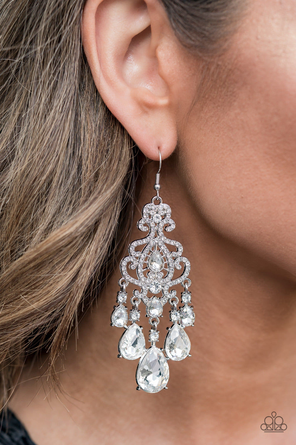 Paparazzi Accessories Queen of All Things Sparkly - White Earrings gradually increasing in size, glassy white teardrop gems create a dramatic fringe at the bottom of a decorative silver frame swirling with dainty white rhinestones for a timelessly over-the-top sparkle. Earring attaches to a standard fishhook fitting.  Sold as one pair of earrings.