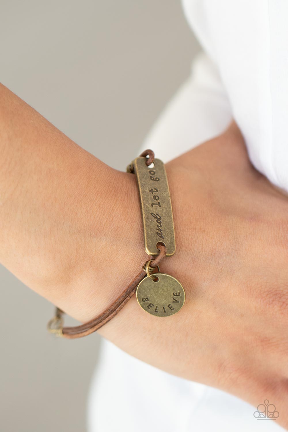 Believe and Let Go - Brass Urban Motivational Bracelets an antiqued disc stamped in the word, "believe" and a brass plate stamped in the phrase, "and let go," are knotted in place around the wrist with layers of brown suede cording, creating a motivational centerpiece. Features an adjustable clasp closure.