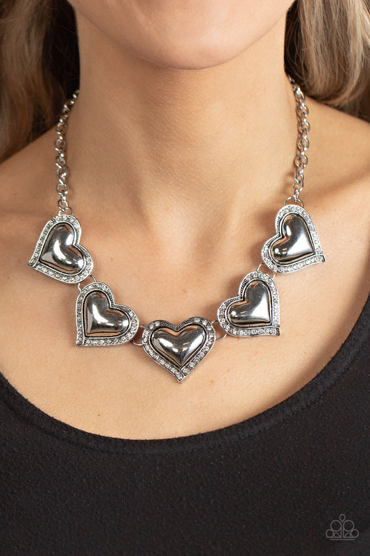 Paparazzi Kindred Hearts - White Heart Necklaces bordered in glassy white rhinestones, an oversized collection of beveled silver heart frames delicately links below the collar for a dramatically romantic look. Features an adjustable clasp closure.  Sold as one individual necklace. Includes one pair of matching earrings.
