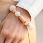 Gold accents and discs combine with a collection of solid, cloudy, and crackle beads in varying sizes and colors to create a modern statement that wraps around the wrist on a stretchy band. The shades of pastel green and soft peach create a fresh and subdued palette, as asymmetrical white pearl beads introduce a hint of refinement.  Sold as one individual bracelet.