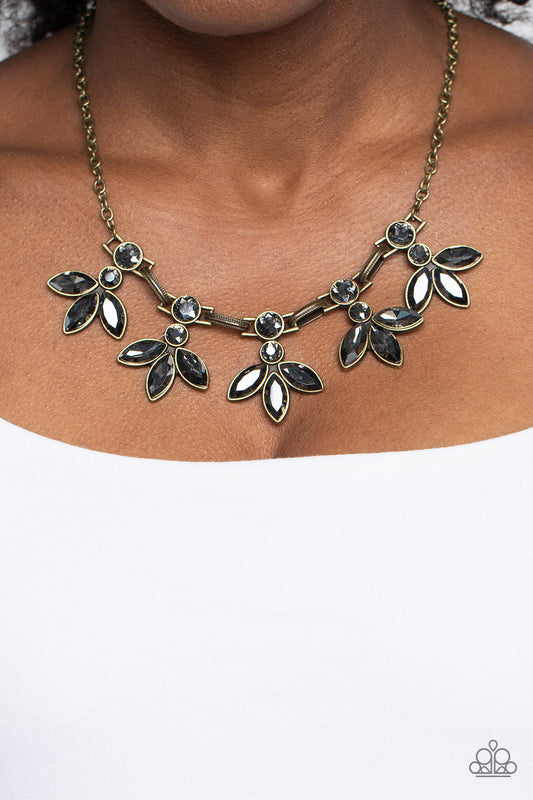 Paparazzi Accessories - Dauntlessly Debonair - Brass EMP 2023 Brass Necklaces featuring smoldering marquise style cuts, oversized smoky gems fan out from an interconnected, circular smoky rhinestone display. Separating the dauntless florals, airy brass ovals, stamped with a dot motif, create additional eye-catching sheen. Features an adjustable clasp closure.  Sold as one individual necklace. Includes one pair of matching earrings.