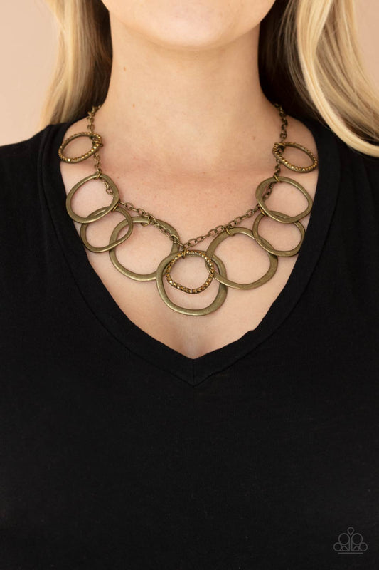 Dizzy With Desire - Brass Circle Link Aurum Rhinestone Necklaces oversized asymmetrical brass rings swing from the bottom of a rustic brass chain. Encrusted in aurum rhinestones, matching brass rings trickle between the dizzying fringe, adding a sparkly smolder to the edgy display. Features an adjustable clasp closure.  Sold as one individual necklace. Includes one pair of matching earrings.  Paparazzi Jewelry is lead and nickel free so it's perfect for sensitive skin too!
